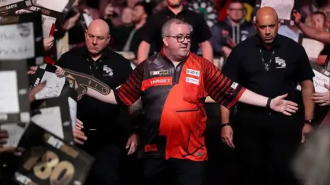 PA Media Stephen Bunting has his arms stretched out towards the crowds who are holding signs. There are three bodyguards behind him as he walks out to start playing. 