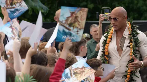 A man with no hair is wearing a button up shirt and sunglasses, speaking to school children holding posters in the air 