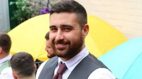 Tom Greest with dark brown hair, and a dark brown beard. He is wearing a suit with a shirt and a brown tie and is smiling. He is standing behind a yellow and blue umbrella. There are three men standing behind him at a distance.