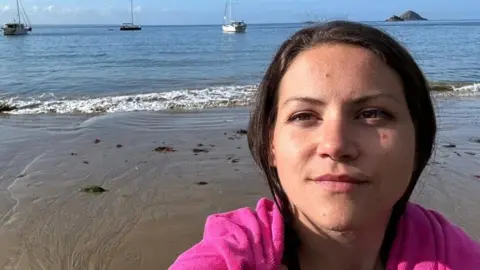 The image shows a woman wearing a pink jumper taking a selfie on a beach. Behind her is wet sand, the sea and three sailing boats in the water. 