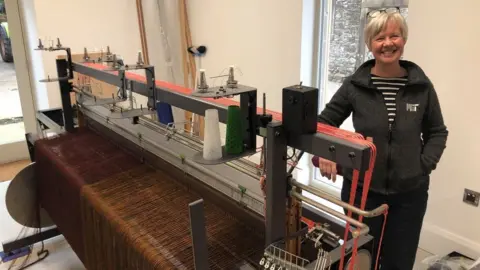 Laura, a woman with short white hair, beams whilst standing next to a pedal loom. It is a large piece of machinery which fills the whole room.