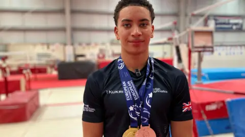 Shariqua Ahmed/BBC Sol Scott with a gold and a bronze medal around his neck, pictured at the gym. He has short black hair and a small moustache, and is wearing a black British Gymnastics branded top.