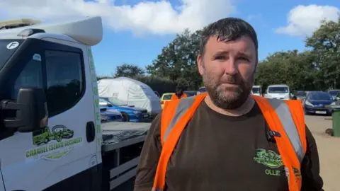 A man with short dark hair and a short dark beard stands outdoors beside a white recovery truck. The person is wearing a black T-shirt and a bright orange high-visibility vest and is facing the camera. The truck door displays the logo and name "Channel Island Recovery", while several parked vehicles, a covered vehicle and trees are visible in the background under a blue sky with scattered clouds.