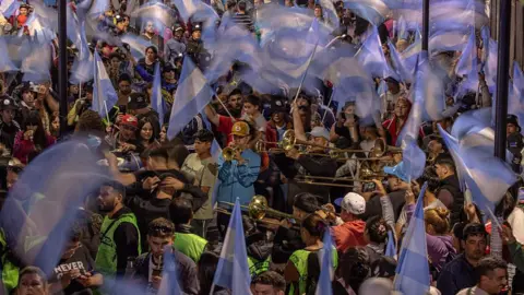 Getty Images People play brass instruments in the middle of a crowd while others wave Argentinian flags