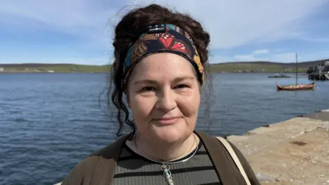 BBC Diane Barnes is a 51-year-old woman with black hair. She is looking at the camera and standing in a harbour area. Sea and hills are behind her and there's a small viking boat to the right. She is wearing a green top, necklace and her her is held up by a bandanna.