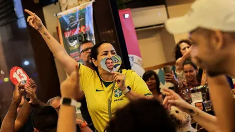 Getty Images People gather around a woman in a yellow Brasil football jersey, raising one hand in the air, as she holds over her mouth a love heart-shaped cutout showing Jair Bolsonaro's face next to the number 22