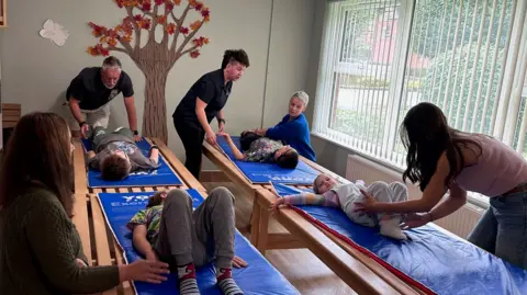 Four children lie on wooden beds topped with blue mats, their parents lean ovver the stretching their legs. A woman with cropped dark hair wearing a Megan Baker House T-shirt is speaking to the children to instruct them in what to do.