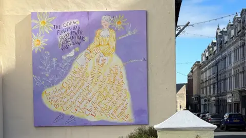 A picture of the mural - it has a purple background, the woman depicted- Josephine, has a long white dress, with Manx words written in gold.