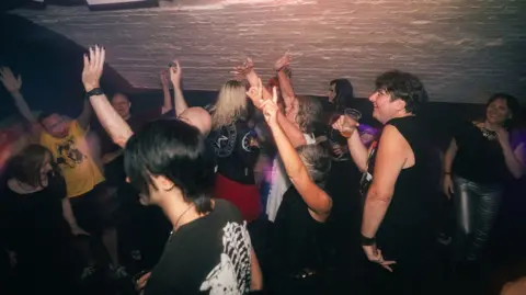 Christopher Illing A group of people dancing inside a building. Some have their hands in the air or are holding drinks. 