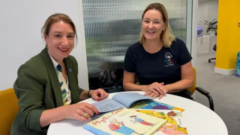 Two women are pictured sitting at a table in front of a pile of books which are open. Both women have their hands on the table and they are both smiling. They appear to be in an office setting. One of the women is wearing a dark green jacket and a white and green shirt and the other woman is wearing a black T-shirt with a logo on one side of it.