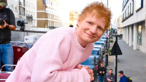 Ed Sheeran smiling smiling and looking at the camera wearing a pink jumper. He is on the top deck of a bus in London