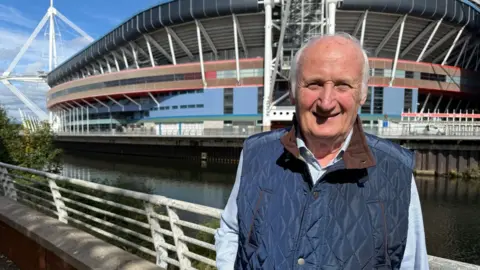A man in a blue Gillet with a shirt underneath. The Principality Stadium can be seen behind him.