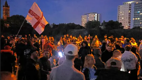 PA Media Demonstrators are standing around, it's quite dark and there are a couple of tall buildings in the background. A red and white flag reads Stop the Boats