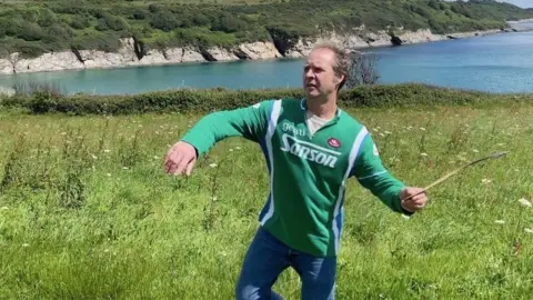 Will Pearson, a man with blonde hair, is holding a wooden boomerang in his left hand and is holding a stance which shows he is about to throw it. He is standing in a green field with wild flowers in it and behind him is the Cornish coast. He is wearing a green sweatshirt and jeans.