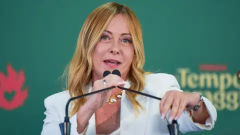 Italian Prime Minister Giorgia Meloni, who has long blonde hair and is wearing a white jacket, speaks into a microphone at an event on 20 September.