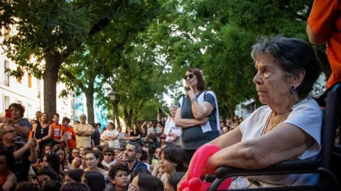 Maricarmen sits outdoors under some trees in a wheelchair. Behind her are a crowd of protesters 