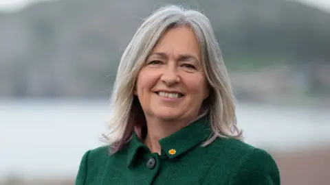 Matthew Horwood/Plaid Cymru Liz Saville Roberts in a green coat smiling with Llandudno in the background, out of focus.