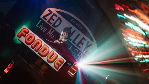 A woman holds a cream telephone to her ear as she stands behind a table with the word "fondue" printed on the front at the Zed Alley Live Music Bar.