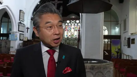 A Japanese man in a suit, with slicked back hair and glasses.