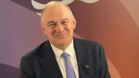Sir Ed Davey, an older man wearing a dark suit, white shirt and patterned lilac tie. He is sat in front of the Liberal Democrats logo, depicting a bird.
