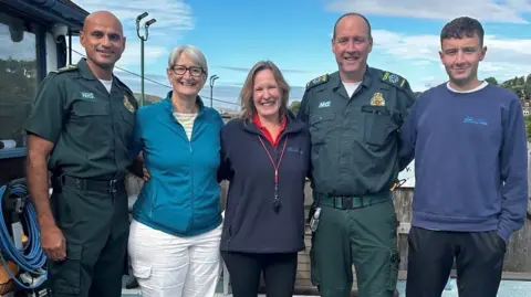 Five people in a row. One man on the far-left in paramedic uniform, next to him is a woman with grey hair, white trousers and a light-blue jacket. In the middle is a woman with shoulder-length light-brown hair wearing a blue fleece with a whistle around her neck. Next to her is a man in paramedic uniform. Next to him is a young man in a blue jumper. 
