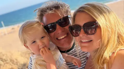 Lily is on a beach with her dad, Jonny, and her mum, Rosie.  The beach and sea are in the background.  Lily is in a sundress; Jonny and Rosie are wearing sunglasses and are smiling at the camera. 
