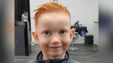Family handout Harrison pictured at hairdressers smiling. He wears a grey zip top and has short red hair styled with gel and a small toy car child's seat is visible behind him.