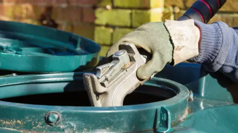 A person's gloved hand holds the end of fuel pipe nozzle as they fill a large, green plastic tank with heating oil.