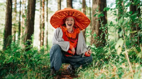 An actor is standing in a woodland setting with a large mushroom hat on as part of a performance. She is smiling at the camera and is holding a magnifying glass in her hand. 