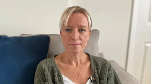 Tracy has blonde hair and is sat on her sofa looking into the camera. She has a white T-Shirt, necklace and green cardigan on.