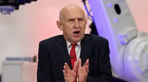 Chancellor John Healey speaking in Coventry in front of purple and grey coloured equipment. He is wearing a dark suit, white short and red tie 