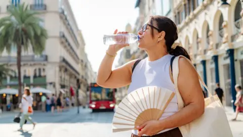 A woman drinks a bottle of watcher with a hand fan in her left hand.