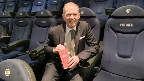 A man in a suit with thinning reddish/blond hair sits on a large cinema seat with empty seats all around him and a tub of popcorn in his hand
