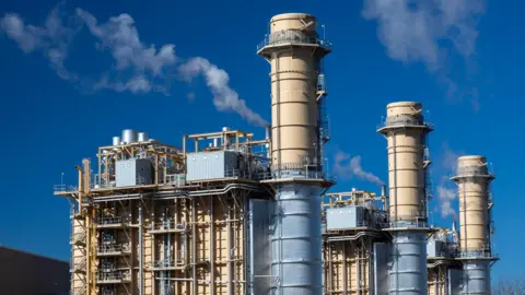 a gas power station with three tall flues. White vapour is seen coming out of some of these.