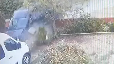 A dark-coloured van crashing through a front wall and into a white van.