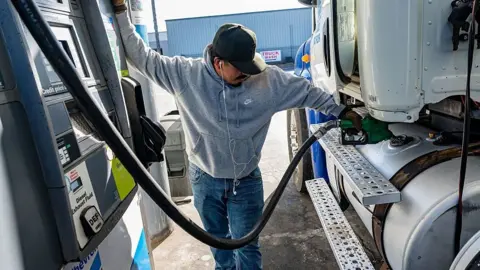 A man, wearing a cap and headphones, looks down as he holds a diesel pump against a lorry 