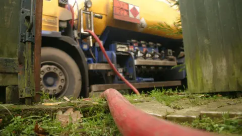 A large red and blue truck parked up delivering domestic heating oil through a red pipe, seen at the front of the shot. 