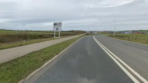 A Google Streetview of the road leading to the planned development, which is green land on the left of the road, and to what would be Seaham Garden Village on the right, where some homes have already been built. The three-lane road leads to a roundabout and is entirely deserted. It's a dry, cloudy day.