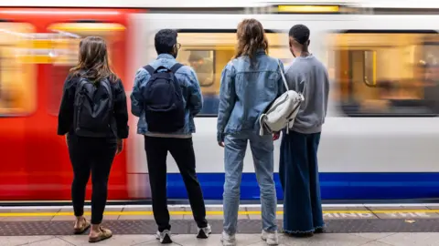 Four young people stand on a London underground platform facing the tracks as a train arrives.