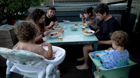 Four friends (two couples) sit at their dinner table outside on the patio with each of their kids in highchairs all eating