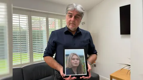 Evanne Miller-Bettridge's father Greg Hughes standing in his living room holding up a framed photograph of his daughter.