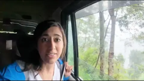 A woman in a blue rain jacket sits in a car, looking out a window.