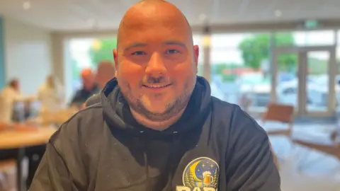 Callum Jarvis is smiling at the camera while sat at a table, with a cafe space blurred out in the background. He has a bald head and black stubble, and is wearing a black hooded top.
