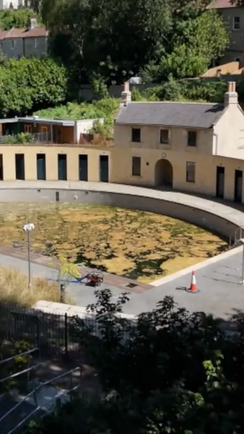 A drone shot of a pool filled with water covered with algae. A Georgian cottage and changing rooms made of Bath stone run along one side of the pool.