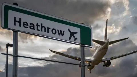 A commercial aeroplane flying low on final approach to land at Heathrow Airport, passing directly over an overhead highway road sign.