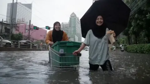 Two women in Thailand navigate thigh-deep floods with a shopping trolley in Thailand