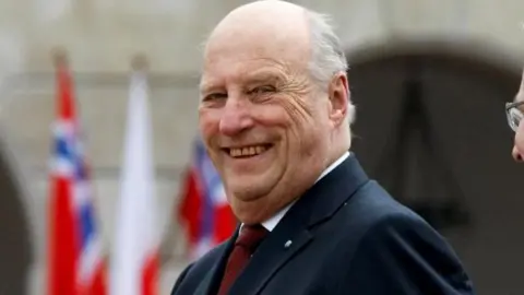 King Harald V smiling to camera with Norwegian flags in background