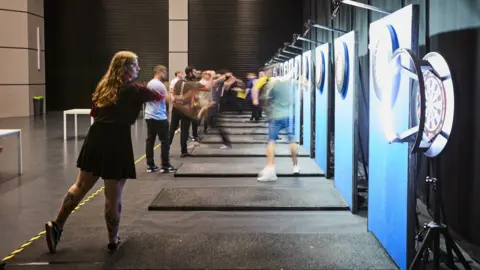 Netflix A row of people throwing darts at dart boards inside a large building.