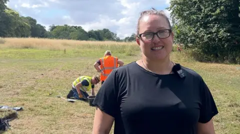 A woman with brown hair, tied back, and black-rimmed glasses smiles as she stands on a large common with green grass and trees. She is wearing a black T-shirt. Behind her, two people in high-vis vests are examining a piece of ground with a bucket standing between them.