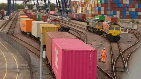 Freight trains with brightly coloured containers at the Port of Felixstowe rail depot, with stacked shipping containers, multiple rail lines and cargo-handling cranes visible in the background.